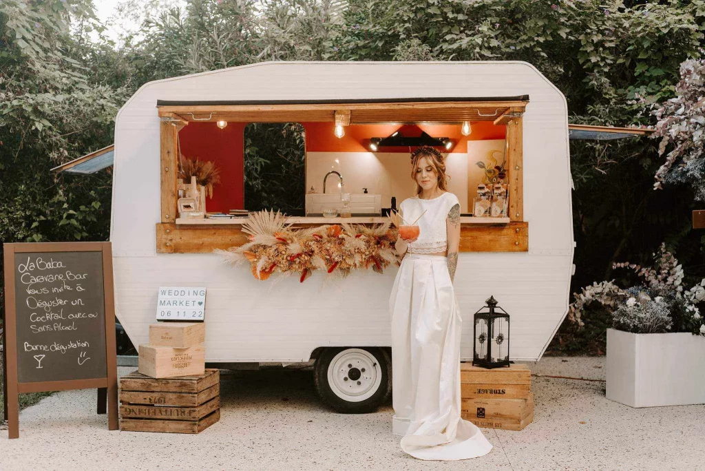 Une caravane aménagée en bar à cocktails, blanche bois et terracota, avec une mariée qui se tient devant entrain de déguster un cocktail dans un verre à gin