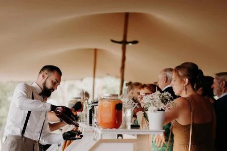 Sous une tente nomade, un barman est entrain de confectionner des cocktails derrière son bar blanc pour les invités du mariage
