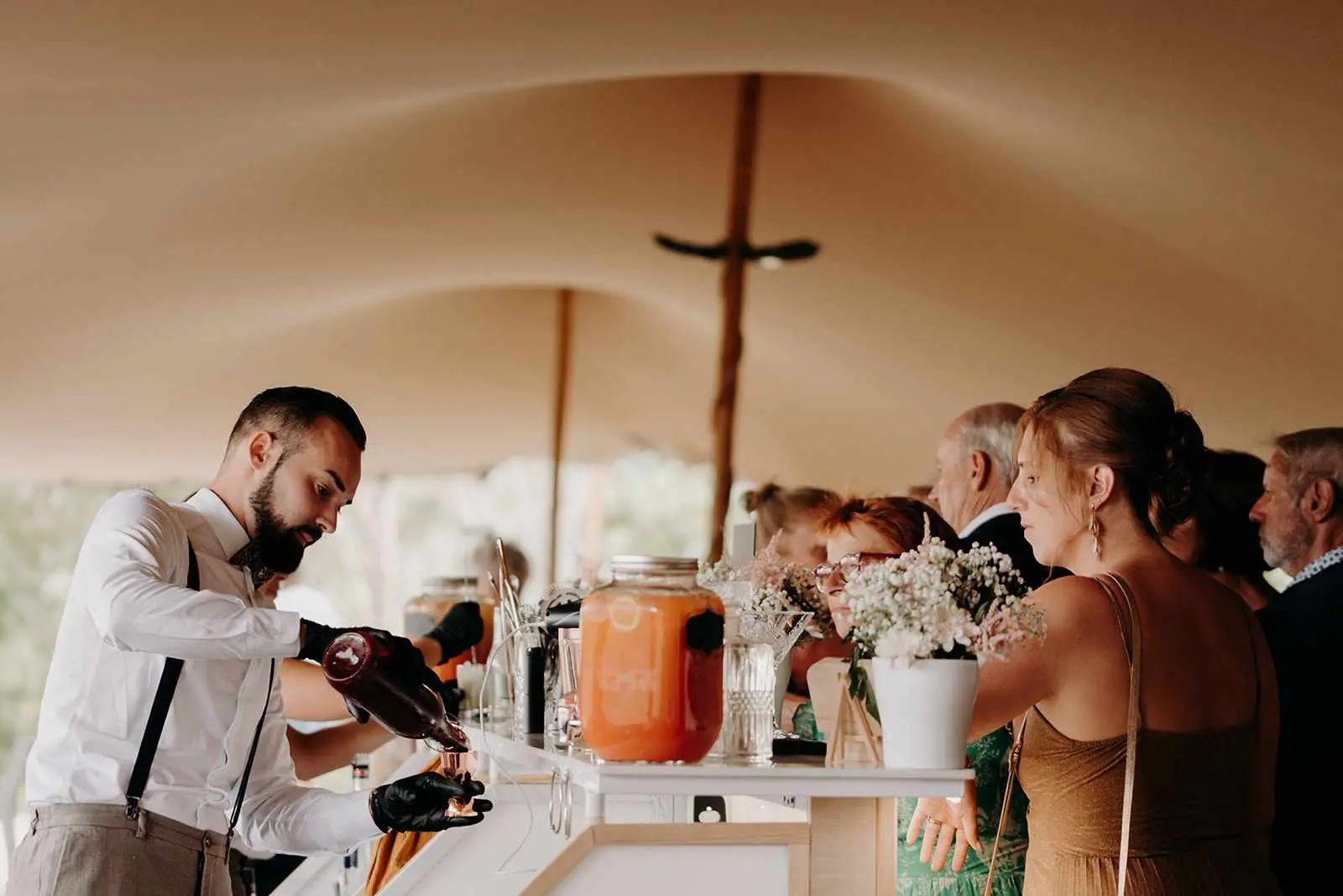 Sous une tente nomade, un barman est entrain de confectionner des cocktails derrière son bar blanc pour les invités du mariage