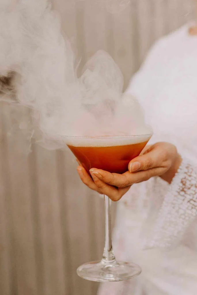 Cocktail rouge rosé dans un verre à martini avec de la fumée au dessus, tenu par une mariée